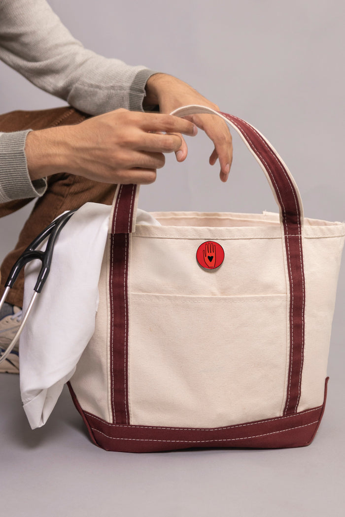 White tote bag with red trim and a red button held by a person on a gray background