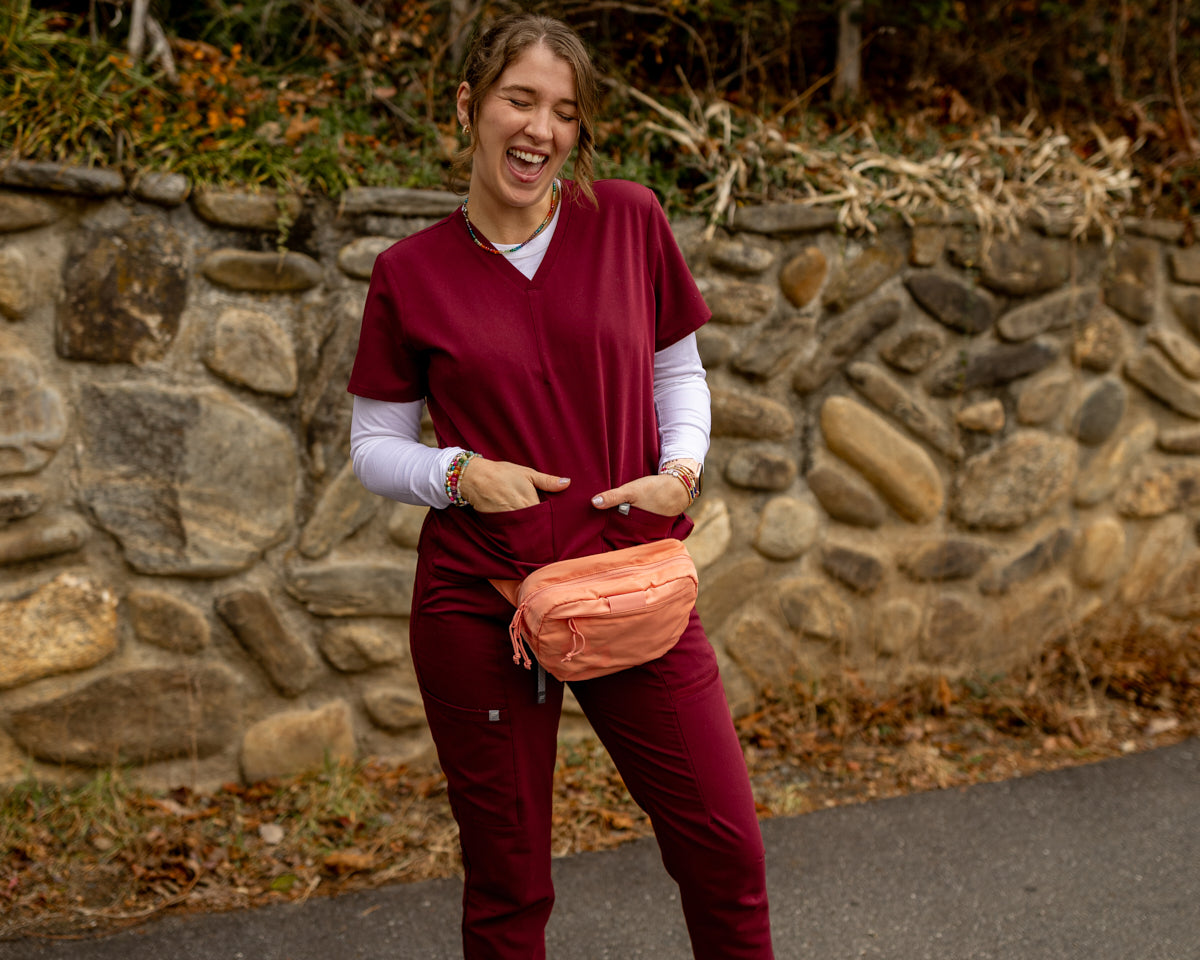 A woman wearing burgundy medical scrubs and a peach fanny pack stands outdoors in front of a stone wall.