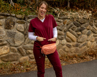 A woman wearing burgundy medical scrubs and a peach fanny pack stands outdoors in front of a stone wall.