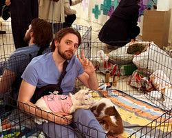 A man in light blue scrubs sits inside a black wire playpen, holding a small white cat wearing a pink shirt. The scene is a busy indoor event with other people and animals visible in the background.