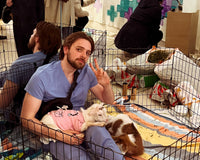 A man in light blue scrubs sits inside a black wire playpen, holding a small white cat wearing a pink shirt. The scene is a busy indoor event with other people and animals visible in the background.