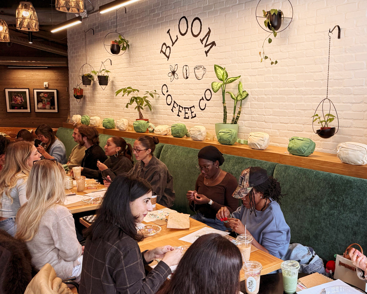 A group of people seated at tables in a cozy cafe interior with a white brick wall featuring the 'BLOOM COFFEE CO' logo. The space is decorated with hanging plants, green velvet booths, and wooden shelves.