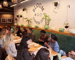 A group of people seated at tables in a cozy cafe interior with a white brick wall featuring the 'BLOOM COFFEE CO' logo. The space is decorated with hanging plants, green velvet booths, and wooden shelves.