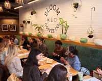 A group of people seated at tables in a cozy cafe interior with a white brick wall featuring the 'BLOOM COFFEE CO' logo. The space is decorated with hanging plants, green velvet booths, and wooden shelves.