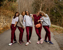 Five women posing together on an outdoor paved path wearing matching athletic tracksuits with white t-shirts and burgundy pants, with a wooded path in the background.