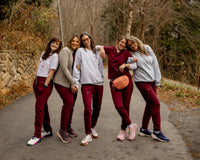 Five women posing together on an outdoor paved path wearing matching athletic tracksuits with white t-shirts and burgundy pants, with a wooded path in the background.
