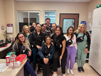 A group of nine healthcare workers wearing matching black and white fanny packs, posing together in a clinical office setting.