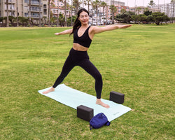 A woman in a black sports bra and leggings performs a yoga pose on a light blue mat in an outdoor park, with two black yoga blocks and a blue bag nearby.